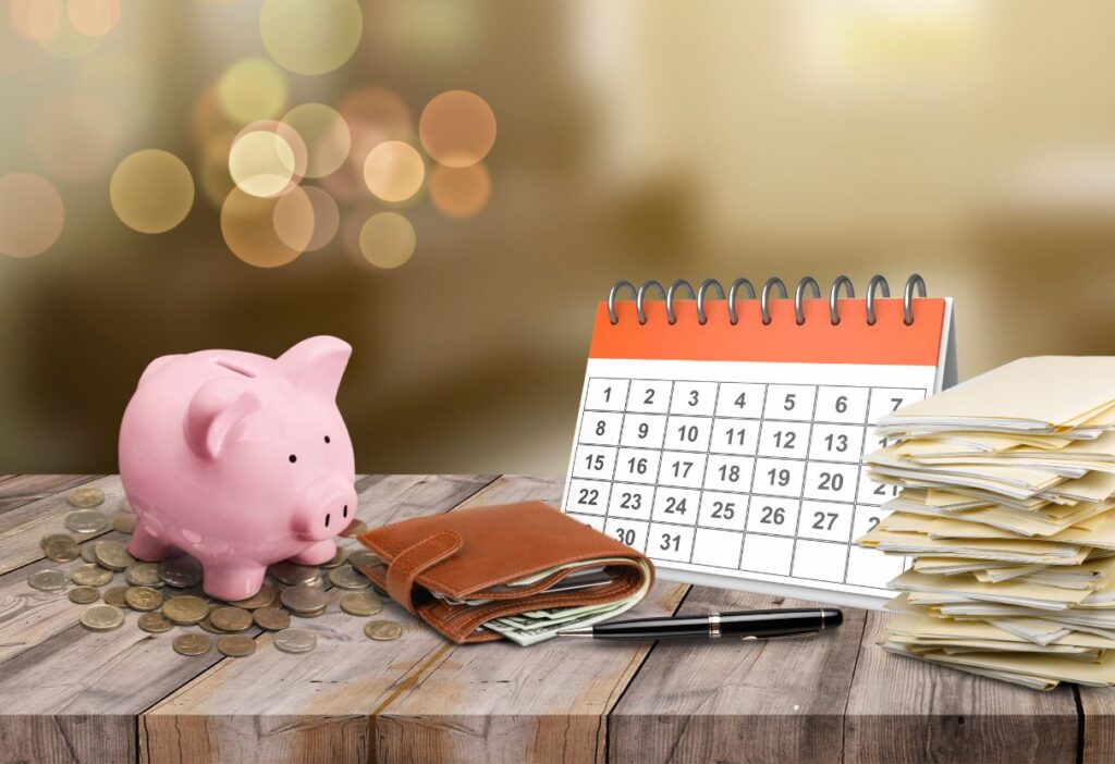 A composition featuring a pink piggy bank surrounded by coins, a leather wallet with cash, a pen, a stack of documents, and a calendar in the background. The image symbolizes financial planning, savings, and organization over time