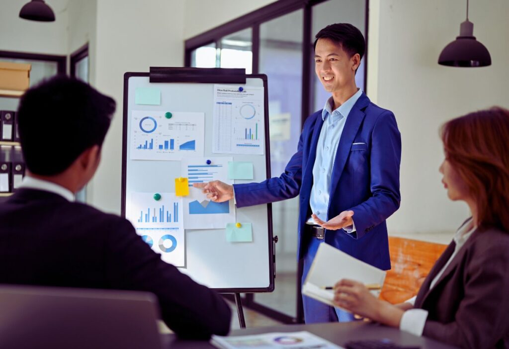 A professional presentation setting with a man in a blue suit pointing at charts and graphs on a whiteboard while explaining to a seated team in an office