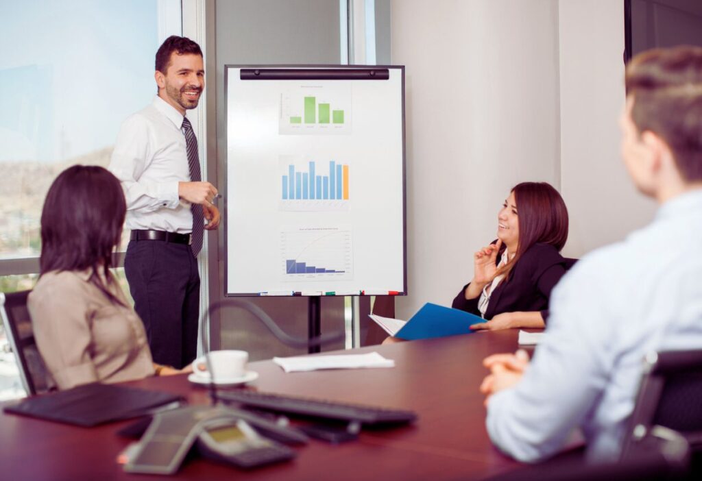 A business meeting scene with a man in a white shirt presenting bar charts and graphs on a whiteboard to a group of colleagues seated in a corporate office