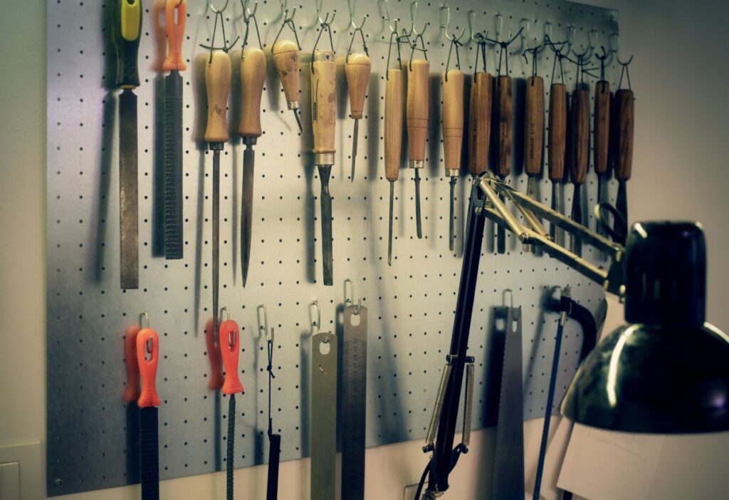 A cluttered workbench with various tools, including a hammer, screwdriver, measuring tape, and paint brushes. A shelf holds jars of nails, screws, and bolts. A toolbox sits open with more tools spilling out