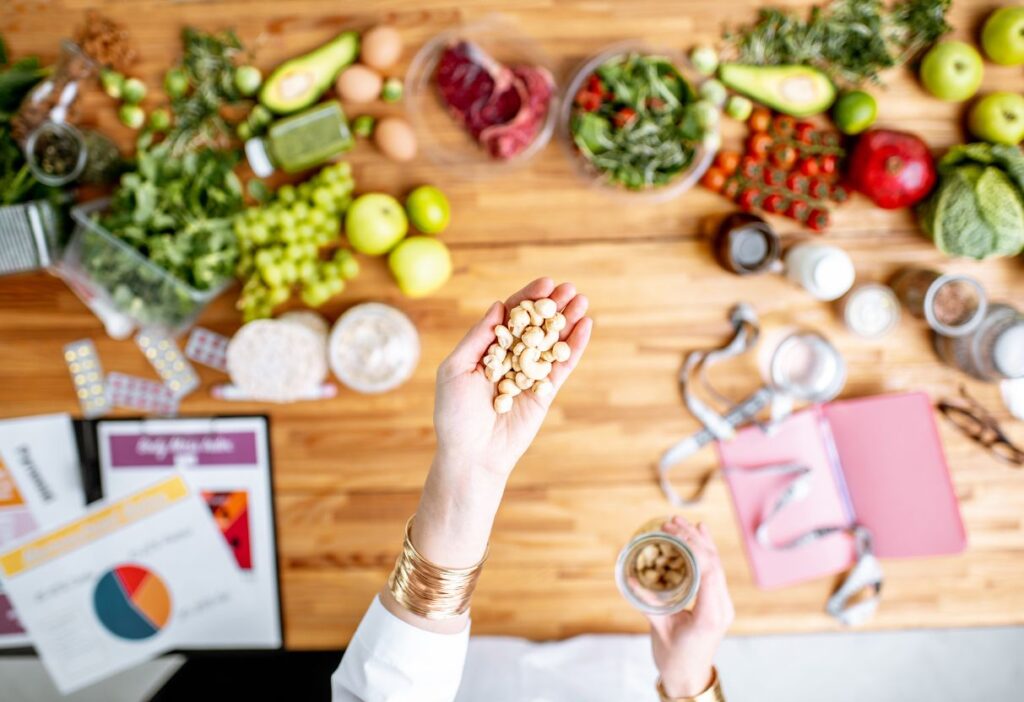 Overhead view of a table filled with fresh fruits, vegetables, and healthy ingredients, including avocados, greens, and nuts. A hand is holding cashews, emphasizing nutrition and mindful eating. The background includes charts and documents, suggesting a focus on dietary planning and health